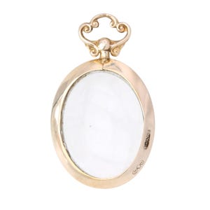 Op de afbeelding: Een goudkleurige ovale hanger met een helder, transparant midden. De hanger heeft een decoratieve lus aan de bovenkant en een kleine markering aan de zijkant. Het ontwerp is eenvoudig en elegant, geschikt voor sieraden.