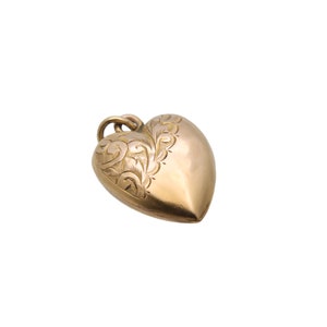 May include: A gold-colored heart-shaped pendant with an ornate, engraved floral design on one side. The pendant has a loop at the top for a chain. The heart is smooth and polished, reflecting light.