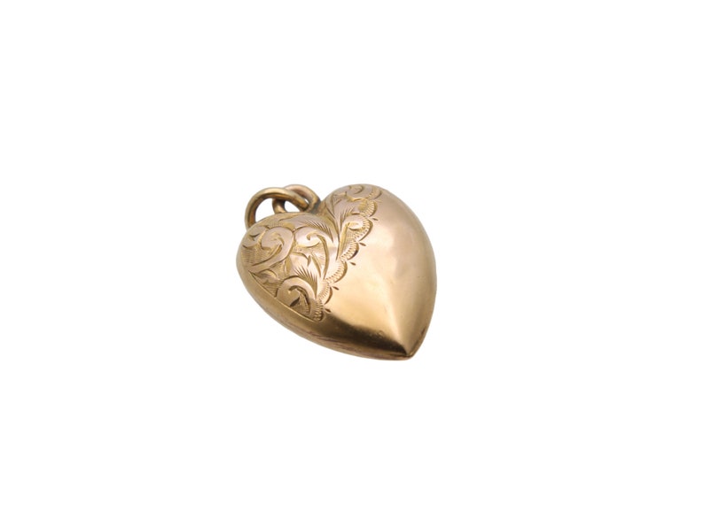 May include: A gold-colored heart-shaped pendant with an ornate, engraved floral design on one side. The pendant has a loop at the top for a chain. The heart is smooth and polished, reflecting light.