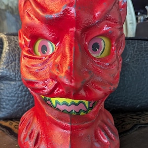 Puede incluir: Figura vintage de cabeza de diablo roja con ojos amarillos y rosas, una amplia sonrisa con dientes verdes y rosas y pequeños cuernos. Decoración de Halloween.