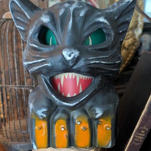 May include: Black cat Halloween decoration with green eyes and a menacing grin showing sharp teeth. The cat has orange paws and sits on a wooden box. A vintage-style piece for Halloween.