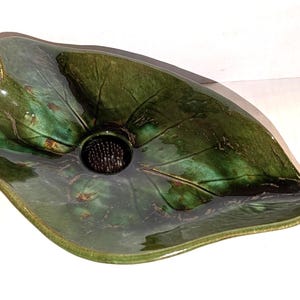 May include: A glossy, dark green ceramic bowl shaped like a large leaf with a prominent central vein and a textured, dark brown circular flower frog insert positioned in the middle of the basin to hold floral stems in place.