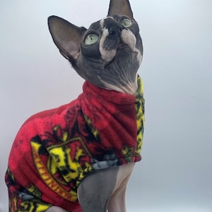 Peut inclure: Un chat sans poil portant un pull en polaire rouge avec un motif jaune et noir. Le pull a un blason de la maison de Poudlard sur le devant.