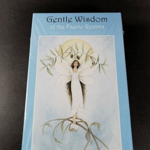 Pode incluir: Uma caixa azul com uma borda branca e o título "Gentle Wisdom of the Faerie Realms" na frente. A caixa apresenta uma ilustração de uma fada com cabelo longo e castanho, asas brancas e um vestido branco, em pé em um galho de uma oliveira. A lua é visível no fundo. O nome do artista, "Sasha St John", está impresso na parte inferior da caixa.