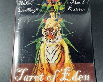 Può includere: Scatola del mazzo di Tarot of Eden con un'illustrazione colorata di una donna che cavalca una tigre con una corona di pappagalli. La scatola è nera con lettere dorate e il testo "Tarot of Eden" e "Urania".