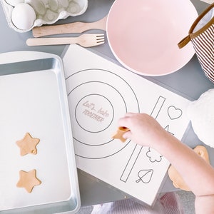 May include: A child's hand places a star-shaped cookie cutter on a white baking sheet with two brown star-shaped cookies. A white baking sheet with a silver rim is on a gray surface. A white bowl with three white eggs is in the background. A pink bowl, a wooden spoon, and a fork are also on the surface. A white sheet of paper with the words "Let's bake together" and a circle with concentric circles is on the surface. A heart, a flower, and a leaf are drawn on the paper.