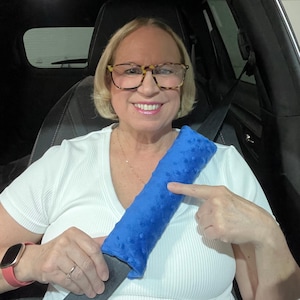 May include: A blue seatbelt cushion with a textured surface. The cushion is being held by a person in a car.