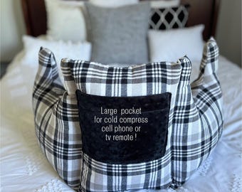 May include: A black and white plaid reading pillow with a black textured pocket on the front. The pocket features the text: Large pocket for cold compress cell phone or tv remote !