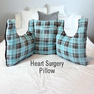 May include: A heart surgery pillow with a blue, gray, and white plaid pattern. The pillow has a pocket and arm supports. The text "Heart Surgery Pillow" is displayed on the front of the pillow.