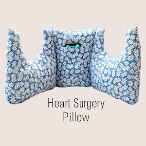 May include: A blue fabric pillow with a white daisy pattern, featuring two side arm supports and a central pocket containing a black smartphone. The text Heart Surgery Pillow is printed in black below the pillow on a light background.