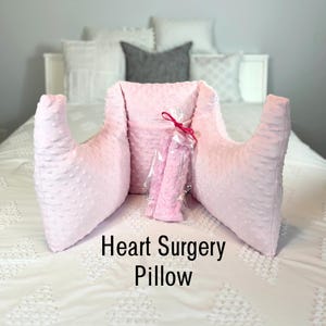 May include: A light pink heart surgery pillow with a textured surface and a matching neck roll, packaged in clear plastic and tied with a red ribbon. The text "Heart Surgery Pillow" is displayed below the pillow.