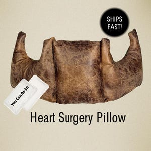 May include: A brown, textured, crescent-shaped heart surgery pillow with a distressed leather-like appearance. A small white rectangular tag attached to the front features the black printed text "You Can Do It!" in a bold, sans-serif font.