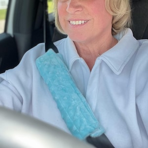 May include: A light blue seat belt cover with a textured surface. The cover is designed to fit over a standard seat belt and is made of a soft material. The seat belt cover is shown in a car.