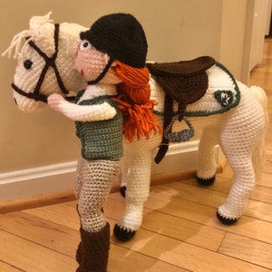 Puede incluir: Un caballo blanco de crochet con una silla de montar marrón y un jinete de crochet que lleva un sombrero de montar marrón y un chaleco verde. El caballo y el jinete están de pie sobre un suelo de madera.
