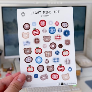 Könnte beinhalten: Ein Stickerbogen mit Knopfdesigns in Rot, Blau, Braun und Weiß. Der Bogen enthält Apfel- und Bärenformen, mit dem Text "LIGHT MIND ART" und "Cozy Fall". Der Bogen misst 10,4 x 14,7 cm.