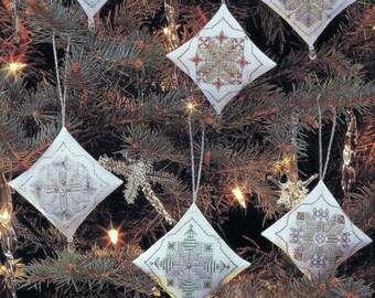 May include: Six white diamond-shaped ornaments with different snowflake designs in silver, gold, and green thread embroidery. The ornaments are hanging on a Christmas tree.
