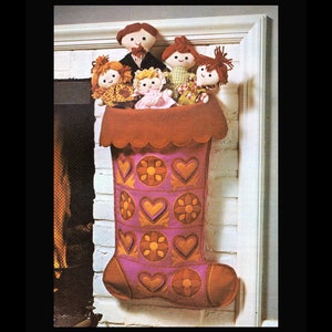 Puede incluir: Una media de Navidad rosa y naranja con un borde festoneado y un diseño de patchwork de corazones y flores. Cinco muñecas asoman por la parte superior de la media.