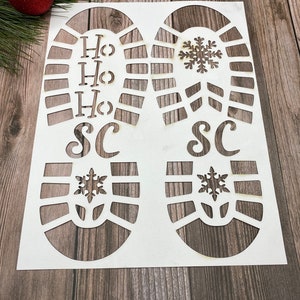 May include: White stencil of two footprints with the words "Ho Ho Ho" and "SC" on one footprint and a snowflake on the other. The stencil is on a wooden surface.