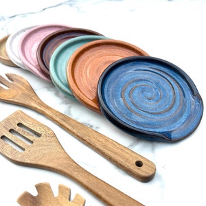 May include: A set of six colorful ceramic spoon rests with a blue spoon rest with a swirl design in the foreground. The spoon rests are arranged in a semi-circle on a white marble surface. Two wooden kitchen utensils are also in the image.