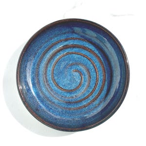 May include: A round ceramic plate featuring a deep blue glaze with a prominent, spiraling copper-toned texture radiating from the center. The edges of the plate are finished with a dark, brownish-black rim that frames the vibrant blue surface.