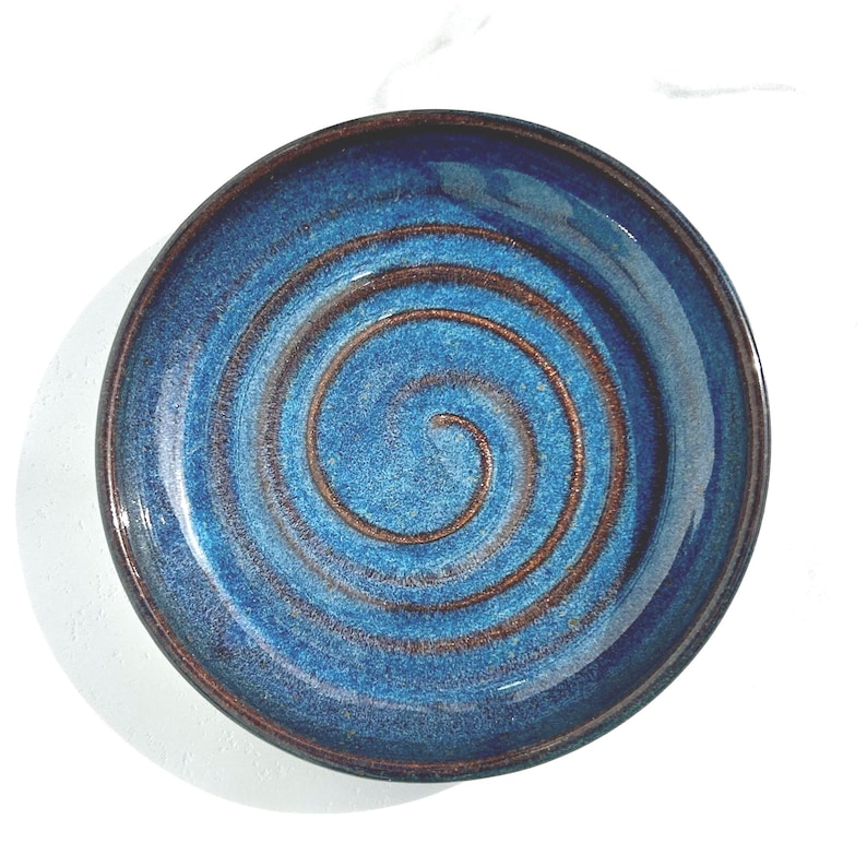 May include: A round ceramic plate featuring a deep blue glaze with a prominent, spiraling copper-toned texture radiating from the center. The edges of the plate are finished with a dark, brownish-black rim that frames the vibrant blue surface.