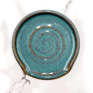 May include: A round ceramic spoon rest featuring a speckled teal glaze with a prominent brown spiral pattern centred in the basin. The edges of the dish are finished with a smooth, dark brown rim, creating a distinct contrast against the blue-green surface.