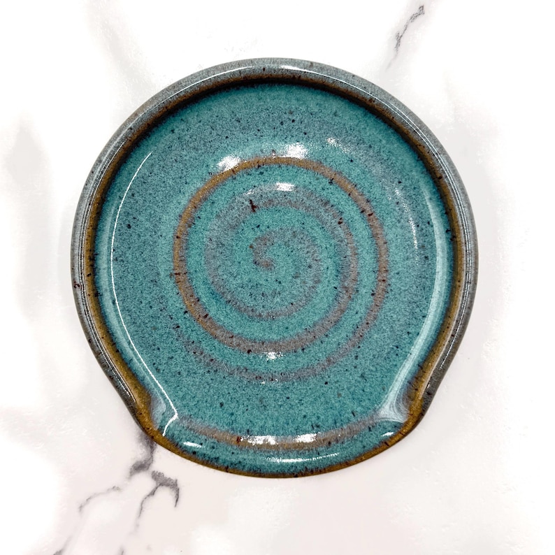 May include: A round ceramic spoon rest featuring a speckled teal glaze with a prominent brown spiral pattern centred in the basin. The edges of the dish are finished with a smooth, dark brown rim, creating a distinct contrast against the blue-green surface.