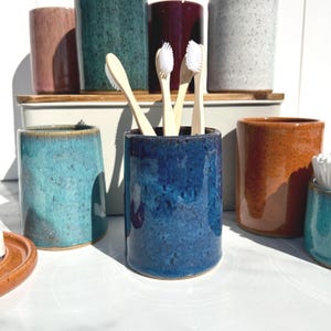 May include: A collection of cylindrical ceramic tumblers in various speckled glazes including blue, teal, orange, pink, and white. The central dark blue tumbler holds three bamboo toothbrushes with white bristles. The items are arranged on a surface.