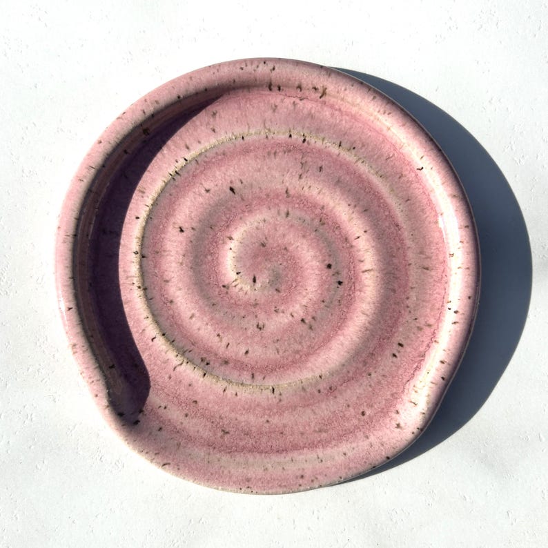 May include: A round ceramic spoon rest featuring a spiral pattern in shades of dusty pink and mauve. The surface has a speckled texture with dark brown flecks throughout, and the edges are slightly raised to contain kitchen utensils during use.