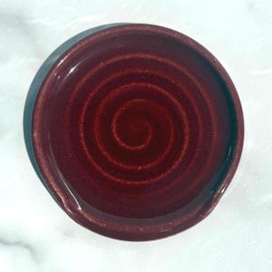 May include: A circular ceramic spoon rest featuring a deep burgundy red glaze with a subtle, swirling spiral pattern embossed into the centre. The dish has a smooth, glossy finish and a slightly raised rim, designed to hold kitchen utensils.