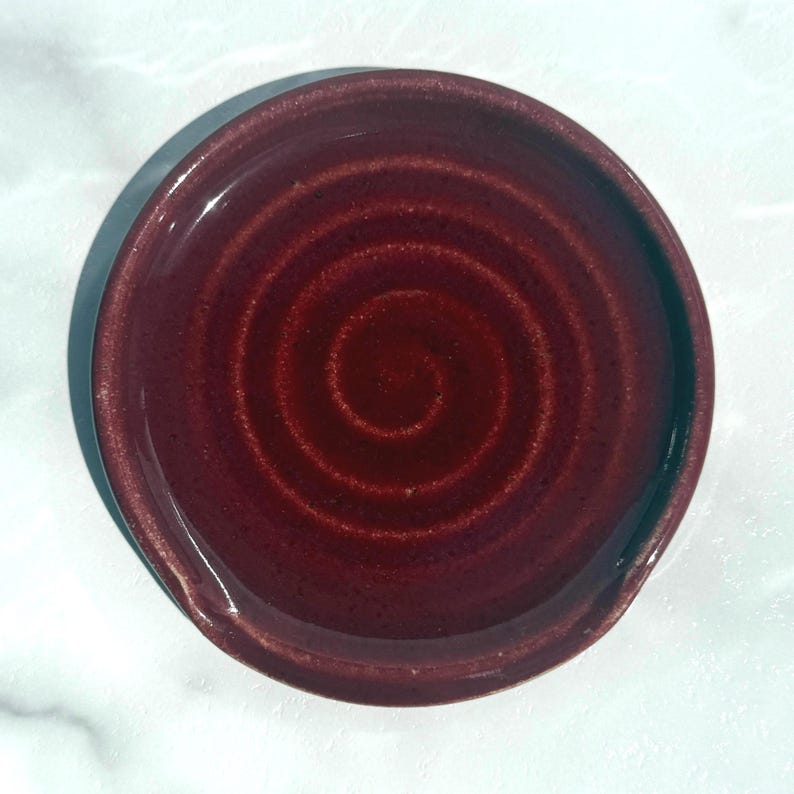 May include: A circular ceramic spoon rest featuring a deep burgundy red glaze with a subtle, swirling spiral pattern embossed into the centre. The dish has a smooth, glossy finish and a slightly raised rim, designed to hold kitchen utensils.