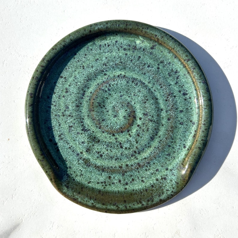 May include: A round ceramic spoon rest featuring a glossy glaze in shades of teal and forest green. The surface displays a dark speckled pattern with a prominent spiral design radiating from the centre towards the raised outer rim of the dish.