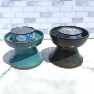May include: Two ceramic elevated pet bowls with a flared base, one in a speckled teal glaze and the other in a dark charcoal glaze. Each bowl holds a silver metal can labeled Great Value Chunk Light Tuna in Water with a blue and white design.