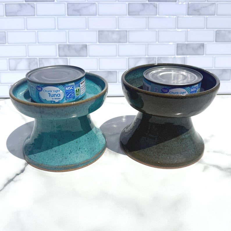 May include: Two ceramic elevated pet bowls with a flared base, one in a speckled teal glaze and the other in a dark charcoal glaze. Each bowl holds a silver metal can labeled Great Value Chunk Light Tuna in Water with a blue and white design.