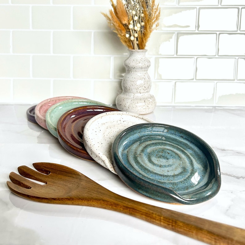 May include: A collection of six round ceramic spoon rests in various colours including blue, white with speckles, dark brown, light green, pink, and dark purple, arranged in a row next to a wooden serving fork and a speckled ceramic vase.