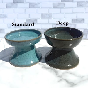 May include: Two ceramic pet bowls with a pedestal base. The left bowl is labeled Standard and has a glossy turquoise glaze. The right bowl is labeled Deep and has a dark, glossy forest green glaze. Both bowls feature a smooth, rounded rim.