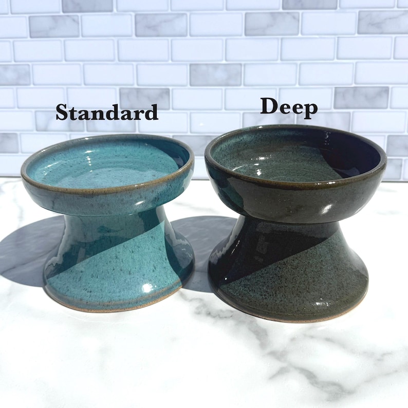 May include: Two ceramic pet bowls with a pedestal base. The left bowl is labeled Standard and has a glossy turquoise glaze. The right bowl is labeled Deep and has a dark, glossy forest green glaze. Both bowls feature a smooth, rounded rim.
