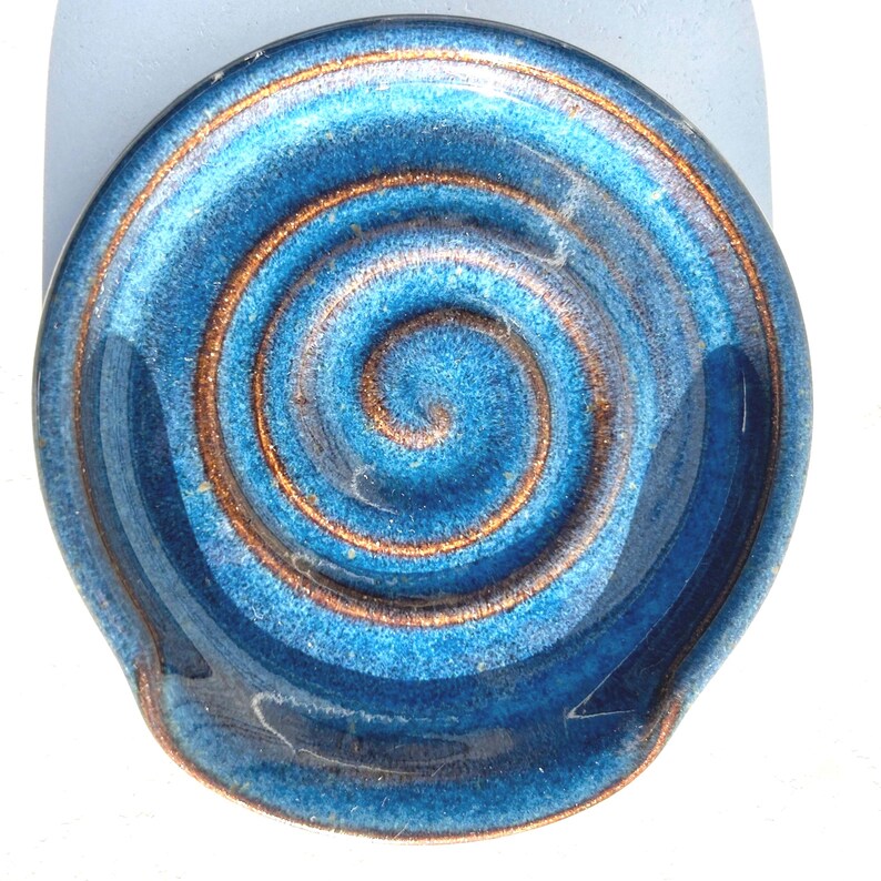 May include: A round ceramic spoon rest featuring a glossy blue glaze with a prominent copper-coloured spiral pattern embossed in the centre. The edges of the dish are slightly raised, and the surface displays a textured finish with deep blue tones.