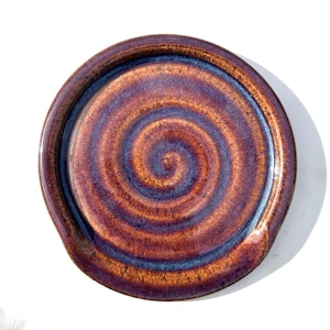 May include: A round ceramic spoon rest featuring a glossy glaze with a swirling pattern in shades of deep copper, burnt orange, and dark blue. The surface has a smooth, reflective finish with a raised outer rim and a slightly indented centre.