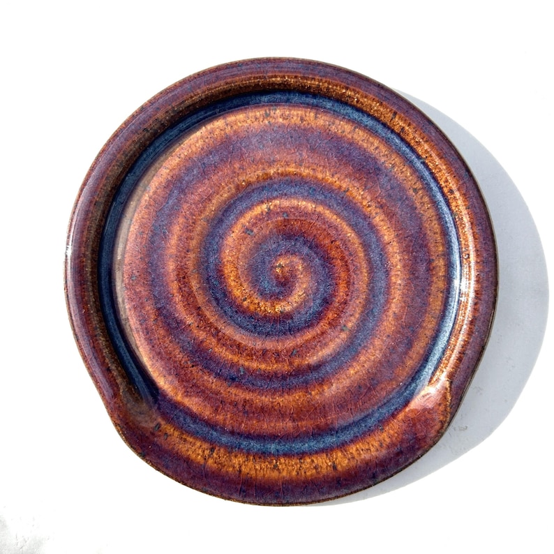 May include: A round ceramic spoon rest featuring a glossy glaze with a swirling pattern in shades of deep copper, burnt orange, and dark blue. The surface has a smooth, reflective finish with a raised outer rim and a slightly indented centre.