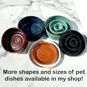 May include: A collection of five ceramic pet dishes in various colors including dark blue, teal, brown, black, and a speckled orange and white bowl. Each dish features a circular spiral pattern on the interior base. Text reads: More shapes and sizes of pet dishes available in my shop!