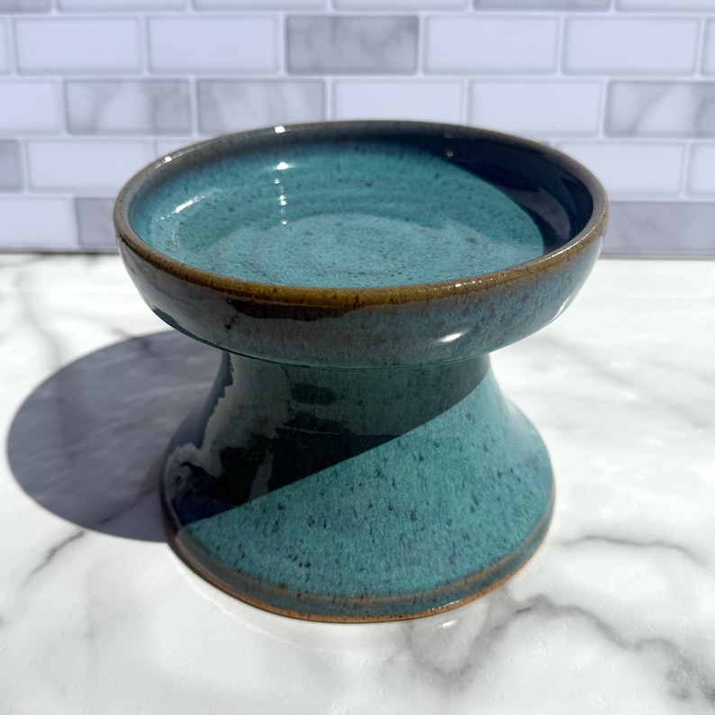 May include: A teal ceramic pedestal bowl with a speckled glaze finish and a dark brown rim. The bowl features a wide, flared base that tapers upward to a circular, shallow basin, showcasing a smooth, glossy texture throughout the entire piece.