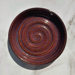 May include: A round ceramic bowl featuring a glossy glaze with a deep red and dark purple spiral pattern radiating from the center. The interior surface displays a textured, swirling design with metallic undertones and a smooth, reflective finish.