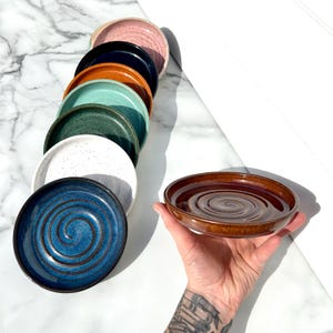 May include: A collection of round ceramic soap dishes in various colors including pink, black, orange, light green, dark green, white, and blue, each featuring a spiral pattern on the interior base, with one brown dish held in the foreground.