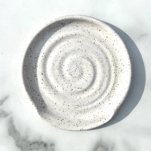 May include: A round ceramic spoon rest featuring a white base with a speckled pattern of small dark brown and black dots. The surface of the spoon rest is moulded with a distinct, raised spiral design that radiates from the centre outwards.