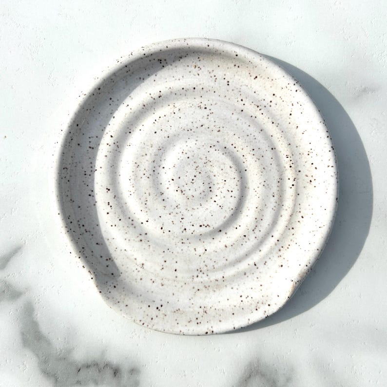 May include: A round ceramic spoon rest featuring a white base with a speckled pattern of small dark brown and black dots. The surface of the spoon rest is moulded with a distinct, raised spiral design that radiates from the centre outwards.