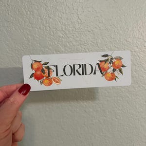 May include: White sticker with an outline of the state of Florida and the word "FLORIDA" in black text. The sticker is decorated with orange blossoms and oranges.