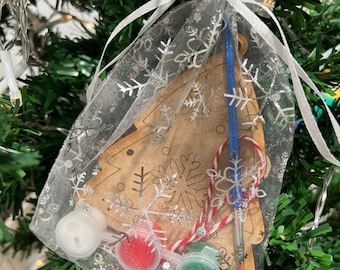 May include: A wooden Christmas tree ornament kit in a white organza bag with silver snowflakes. The kit includes three small paint pots, a paintbrush, and a red and white candy cane.