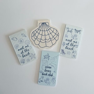 May include: Four bookmarks with nautical themes. Two rectangular bookmarks feature blue stripes and the text meet me at the beach. One rectangular bookmark features marine life and the text ocean lovers book club. One die-cut bookmark is a seashell.
