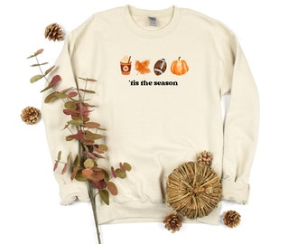 Puede incluir: Una sudadera de color crema con un gráfico de una taza de café, una hoja, una pelota de fútbol y una calabaza. El texto "'tis the season" está impreso debajo del gráfico.